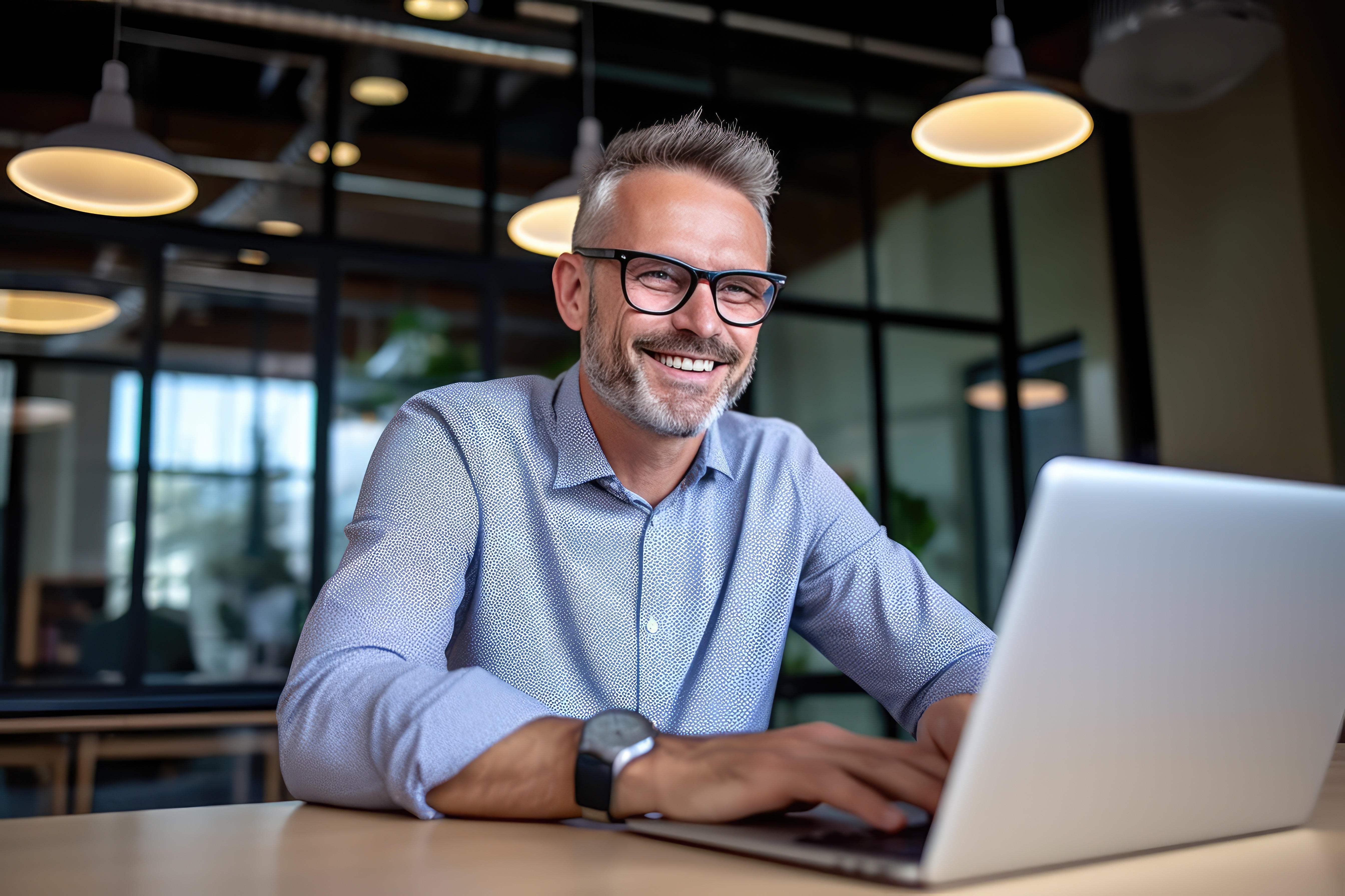 Smailing man in a button down shirt typing on a laptop at a table